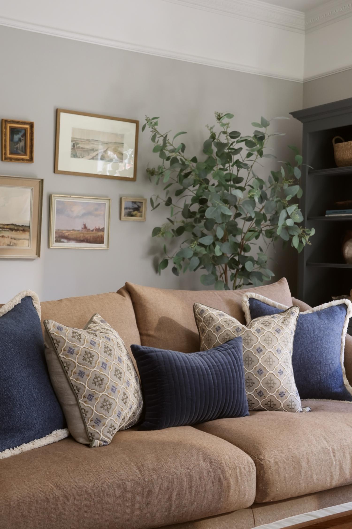 Living room with a beige sofa adorned with blue and patterned cushions, framed pictures on the wall, and a plant.