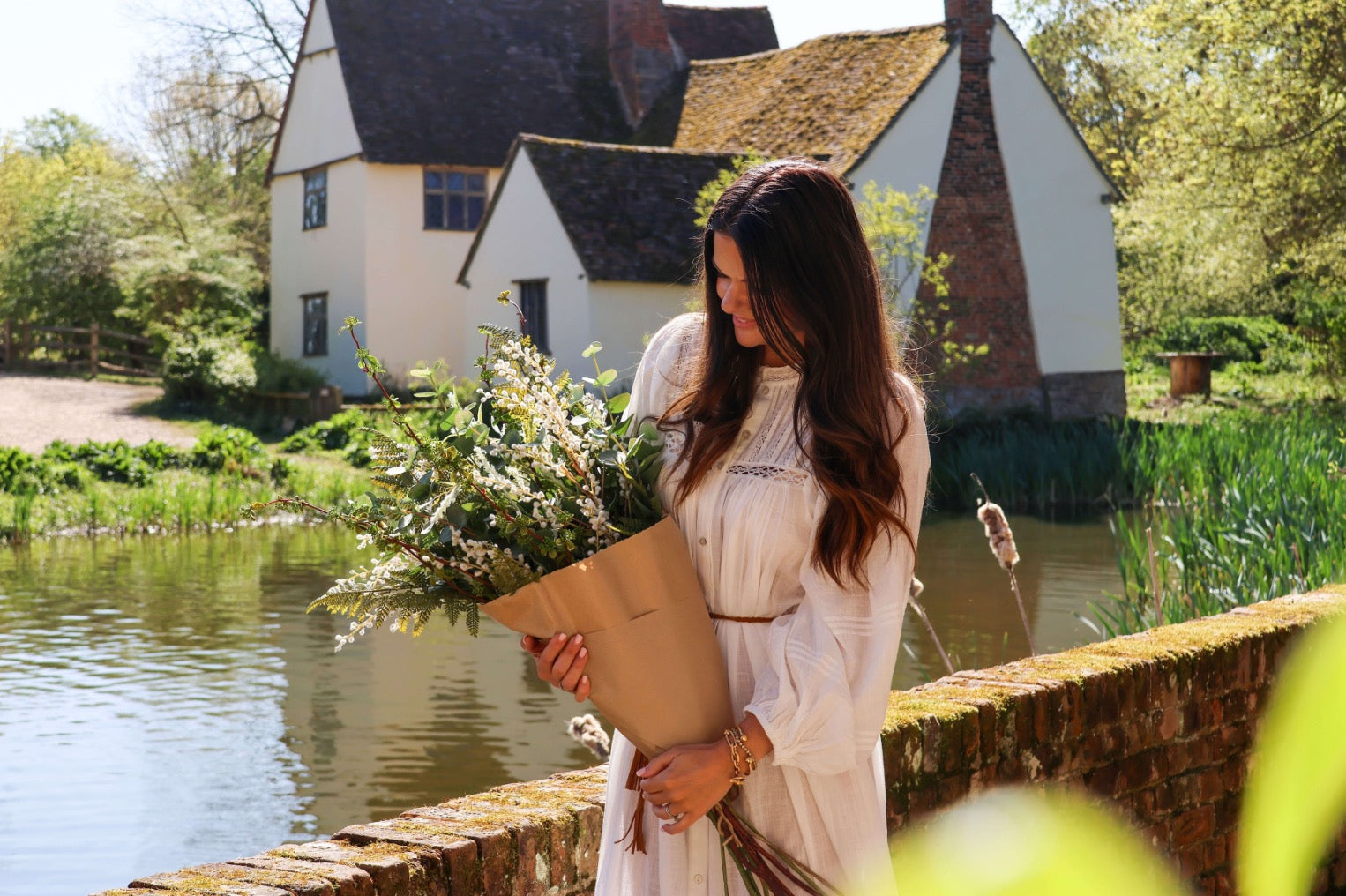 Woman holding a faux green foliage bouquet by a pond with a traditional house in the background