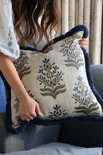 Person holding a decorative cushion with floral embroidery and navy blue fringe trim.