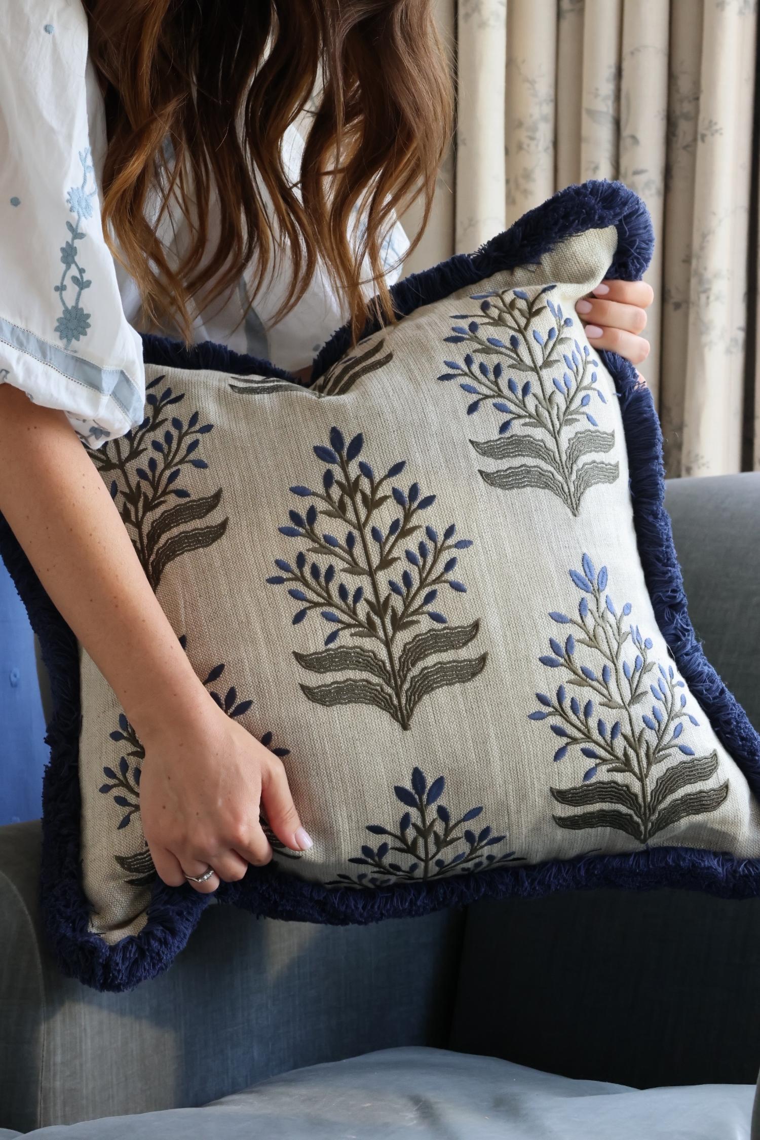 Person holding a decorative cushion with floral embroidery and navy blue fringe trim.