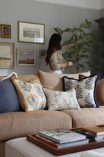 Woman arranging a faux tree next to a brown sofa with decorative cushions in a living room.