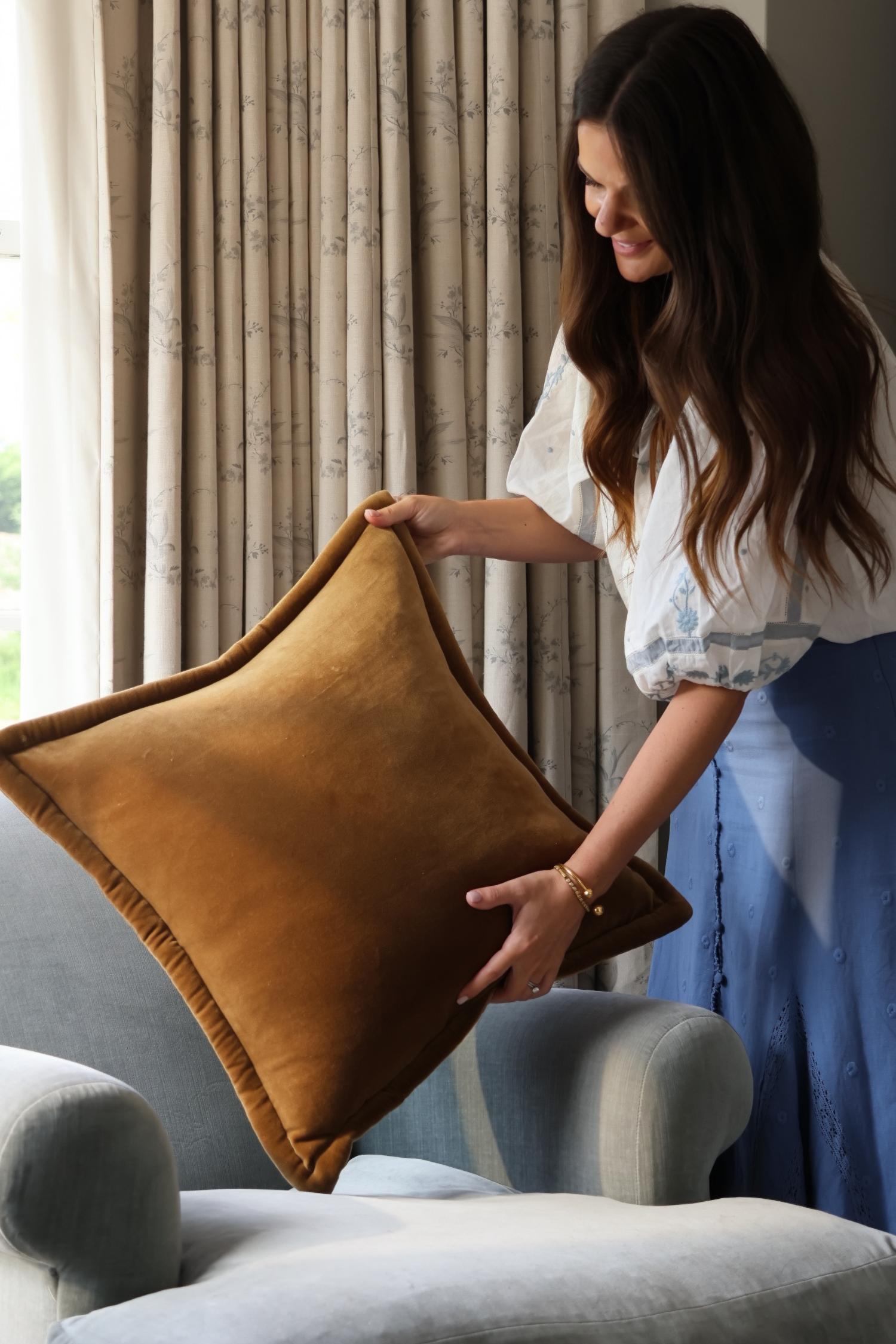 Woman arranging a brown pillow on a gray sofa in a room with curtains.