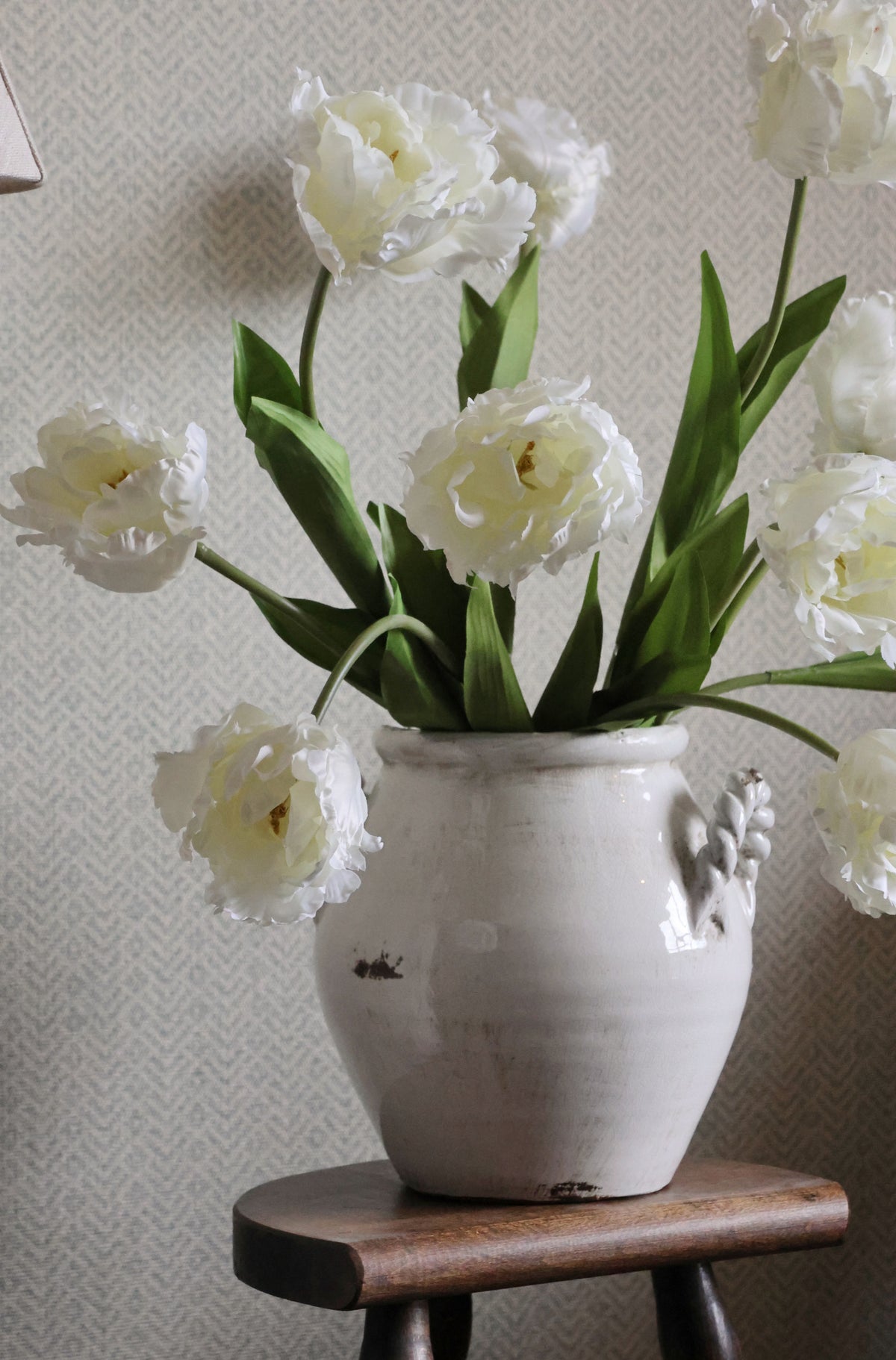 White double tulips in a decorative vase on a wooden stool against a light gray wall.