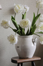 White double tulips in a decorative vase on a wooden stool against a light gray wall.