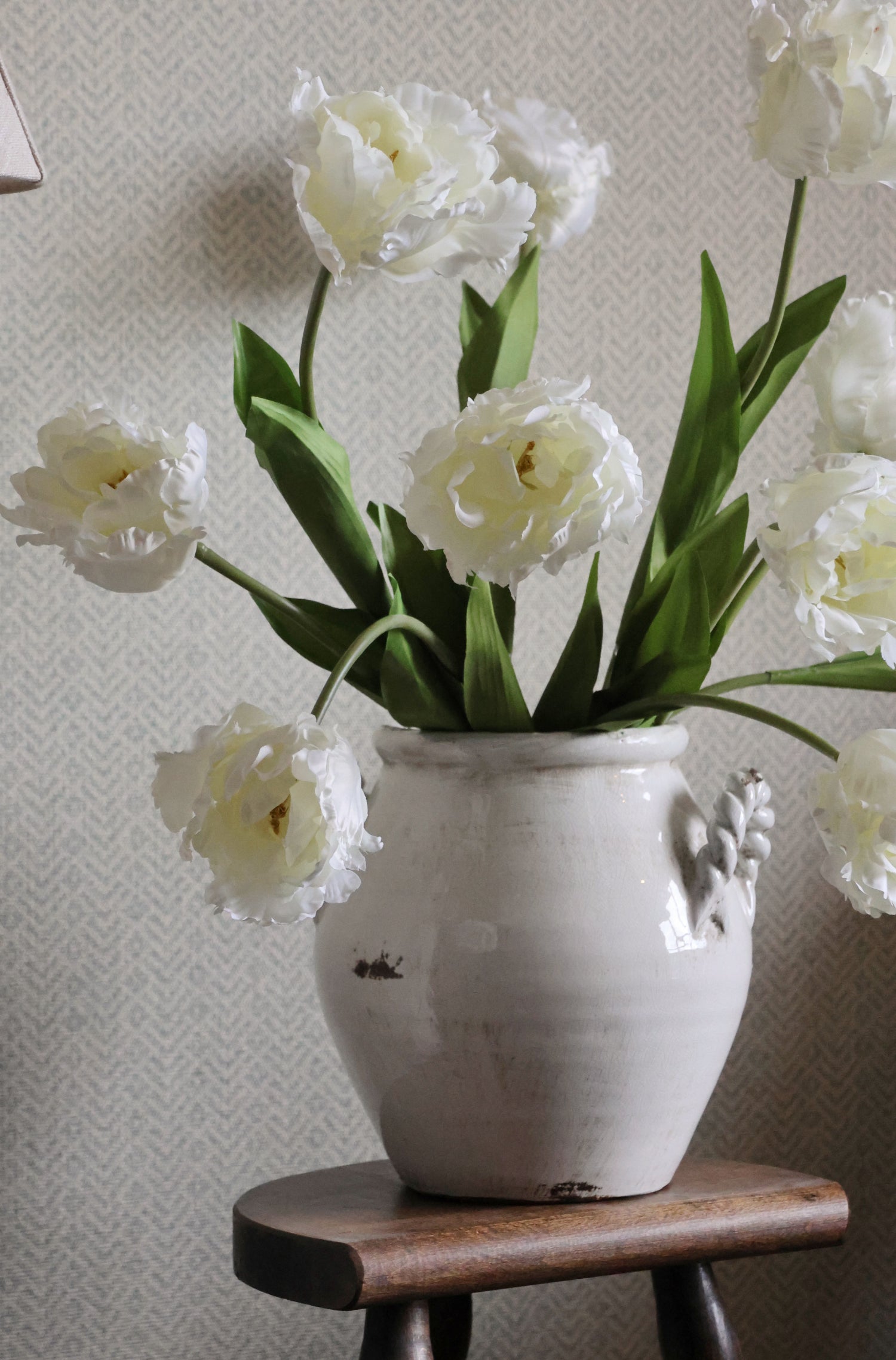 White double tulips in a decorative vase on a wooden stool against a light gray wall.
