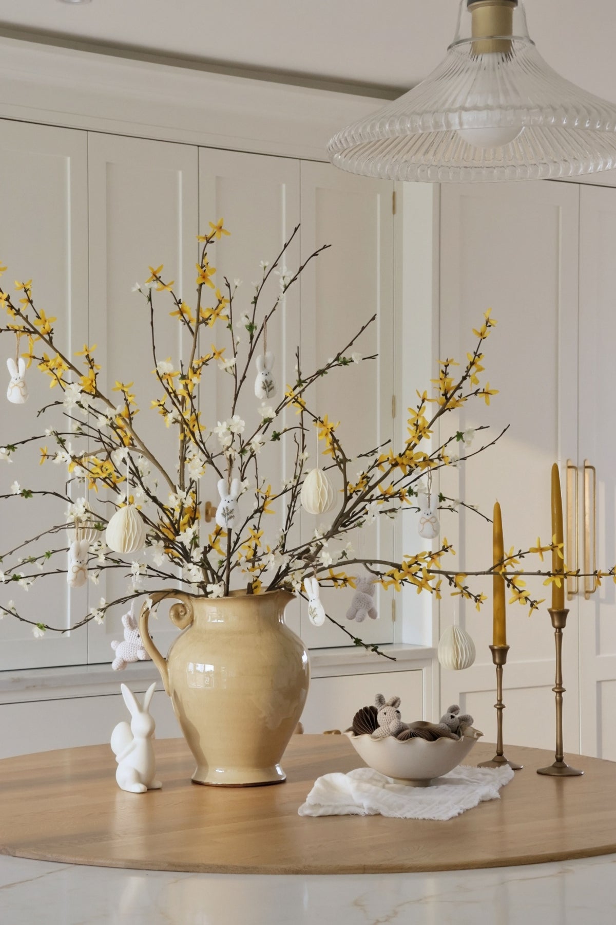 Decorative table setting with a vase of branches, candles, and small figurines in a room.