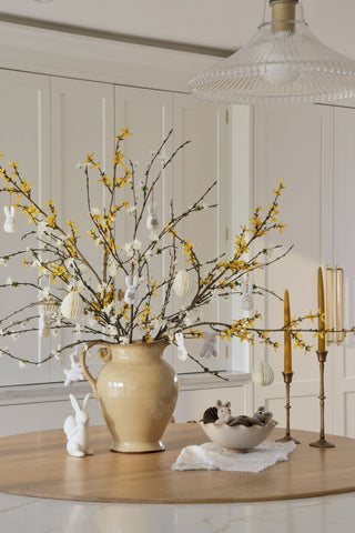 Decorative table setting with a vase of branches, candles, and small figurines in a room.