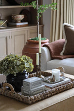 A woven rectangular tray styled in a cozy living room setting, holding a black vase with green hydrangeas, a stack of gray decorative boxes, silver candlesticks, and a white candle.