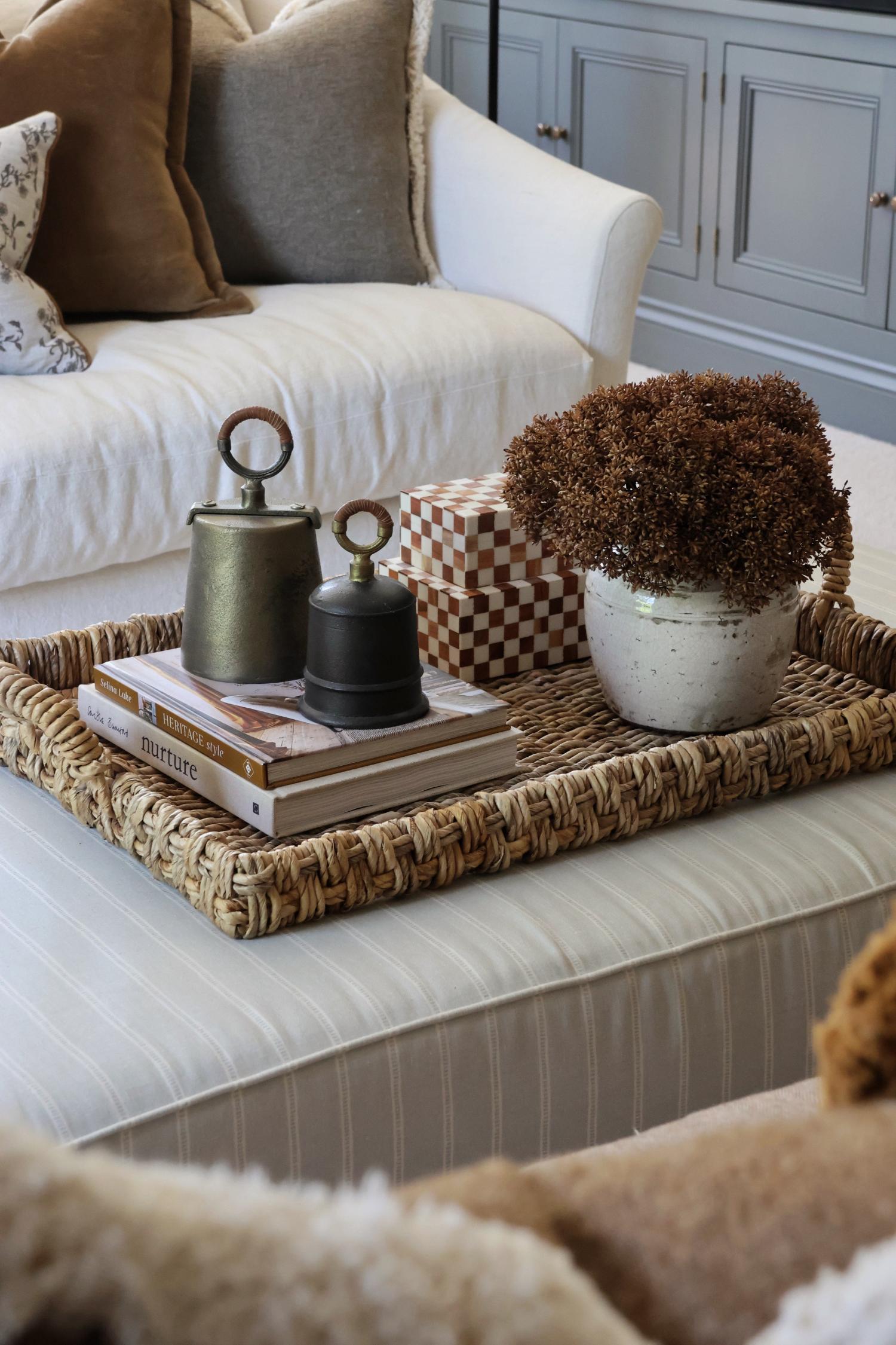 Large woven tray on a footstool styled with antiwue brass bells, a short white vase with brown artificial stems and checkered resin boxes. 