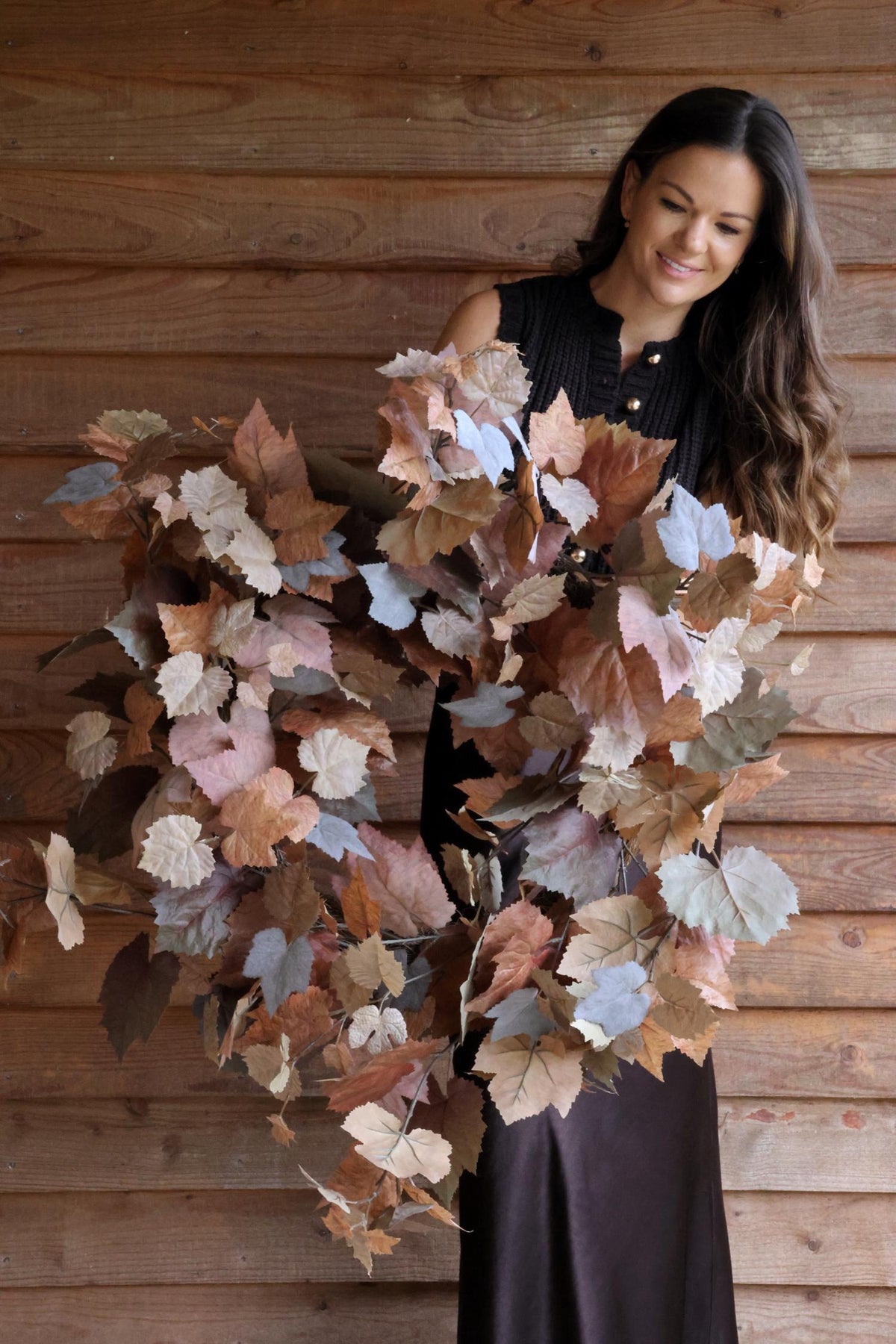 Woman holding a large artificial autumn wreath with maple leaves against a wooden wall