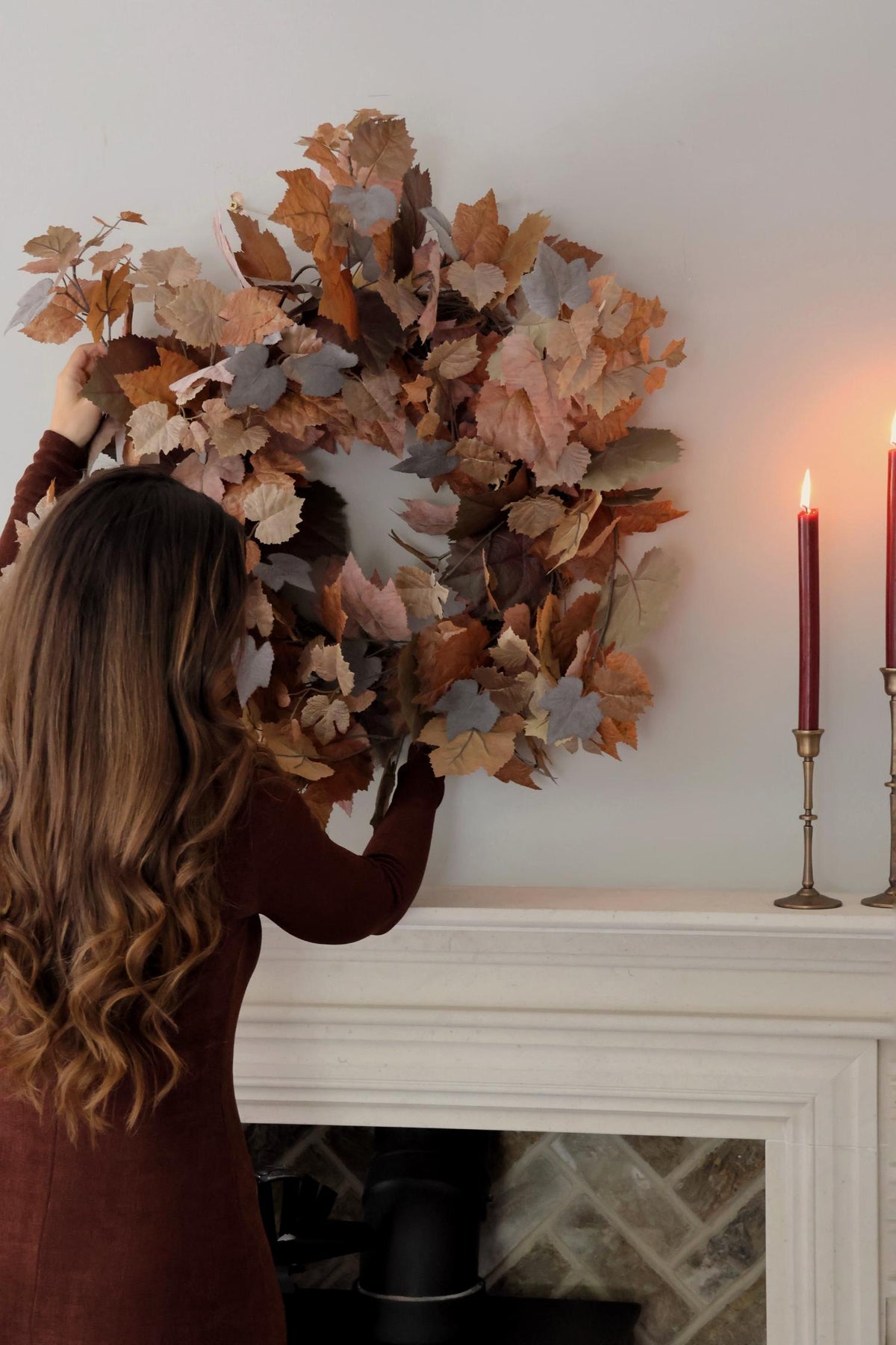 Person adjusting an artificial autumn leaf wreath on a fireplace mantel with candles in the background