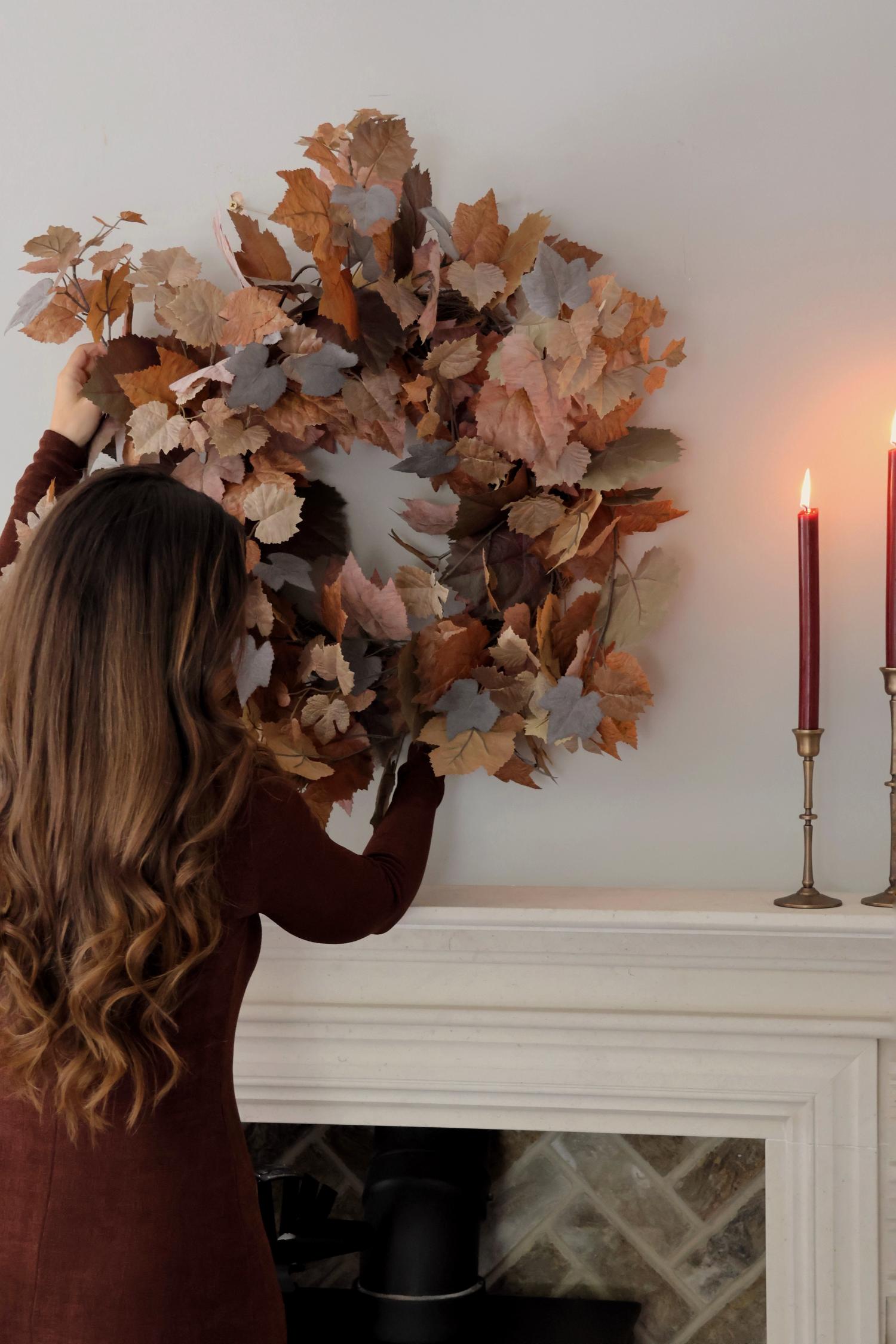 Person adjusting an artificial autumn leaf wreath on a fireplace mantel with candles in the background