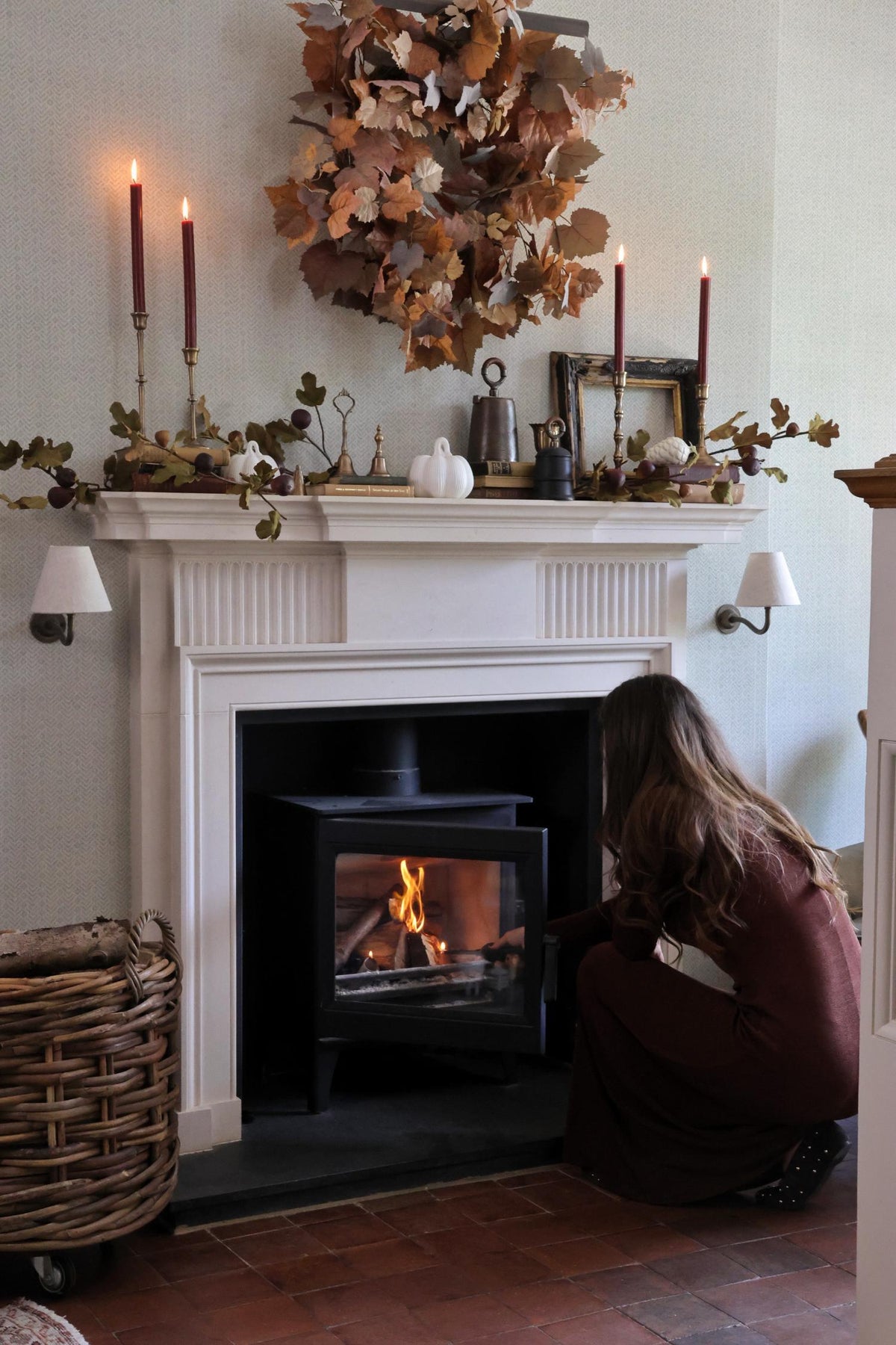 Woman sitting by a  cosy lit logburner with an autumn decorated mantel and candles.