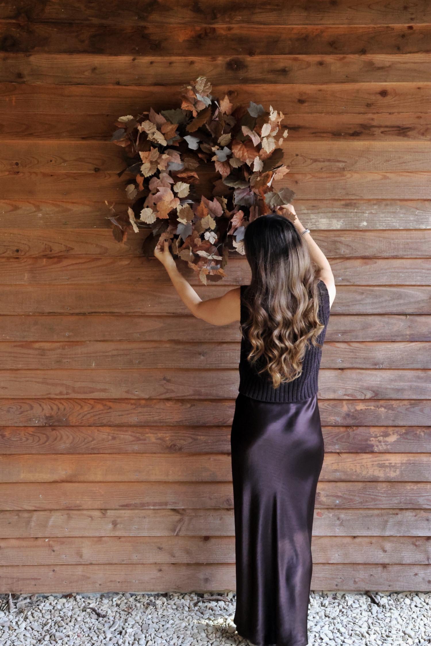 Woman in a long dark dress holding a large artificial autumn wreath against a wooden wall.