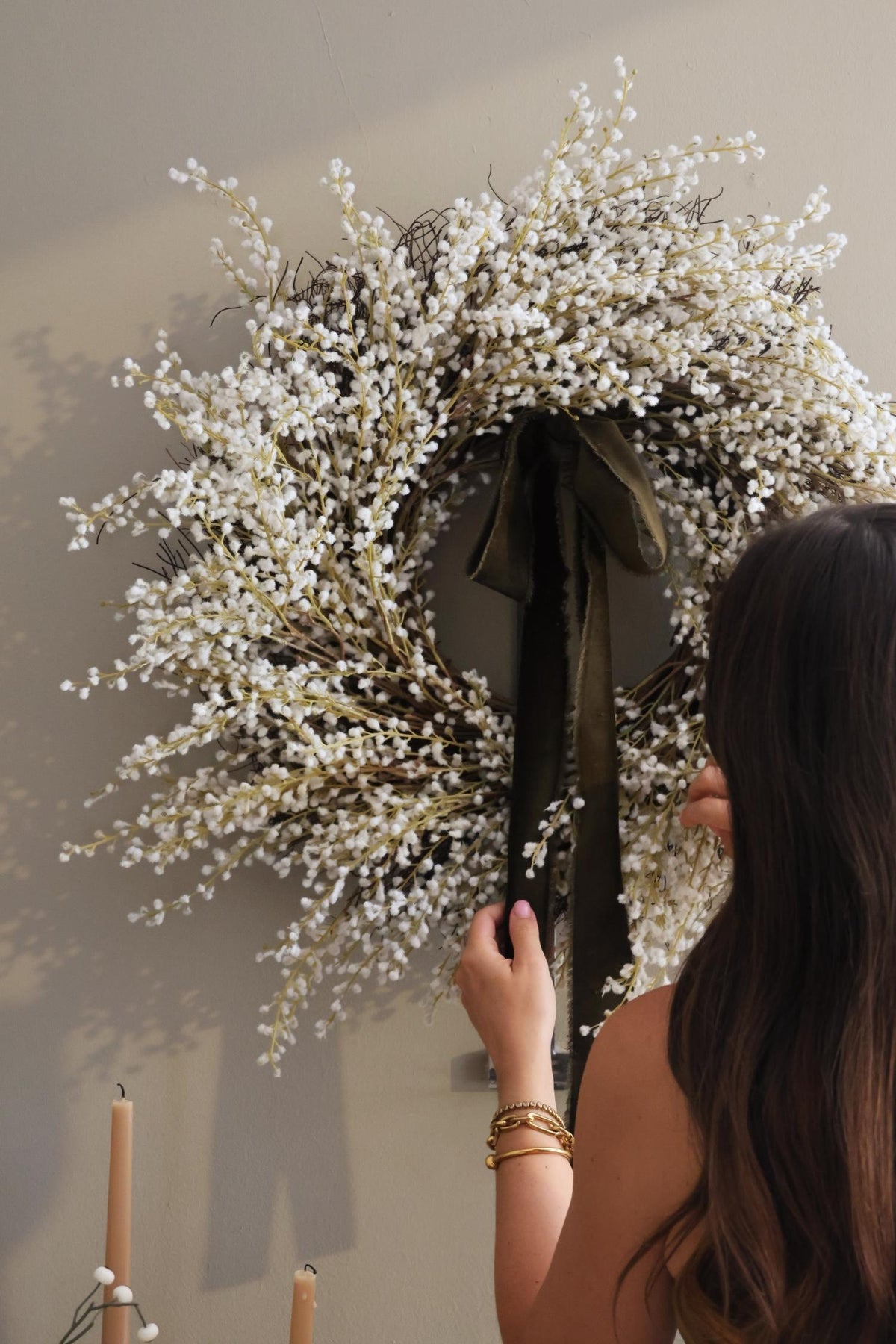 women arranging large artificial white willow wreath with dark green ribbon on a plain wall 