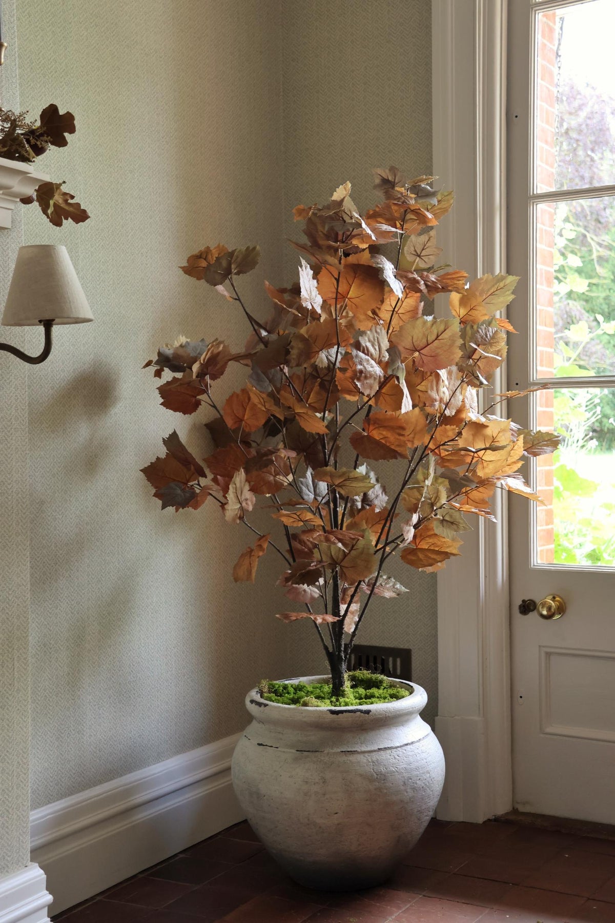 An artificial autumn leafy tree with brown and orange leaves in a textured gray pot, placed in a corner of a room with beige walls and a white door.
