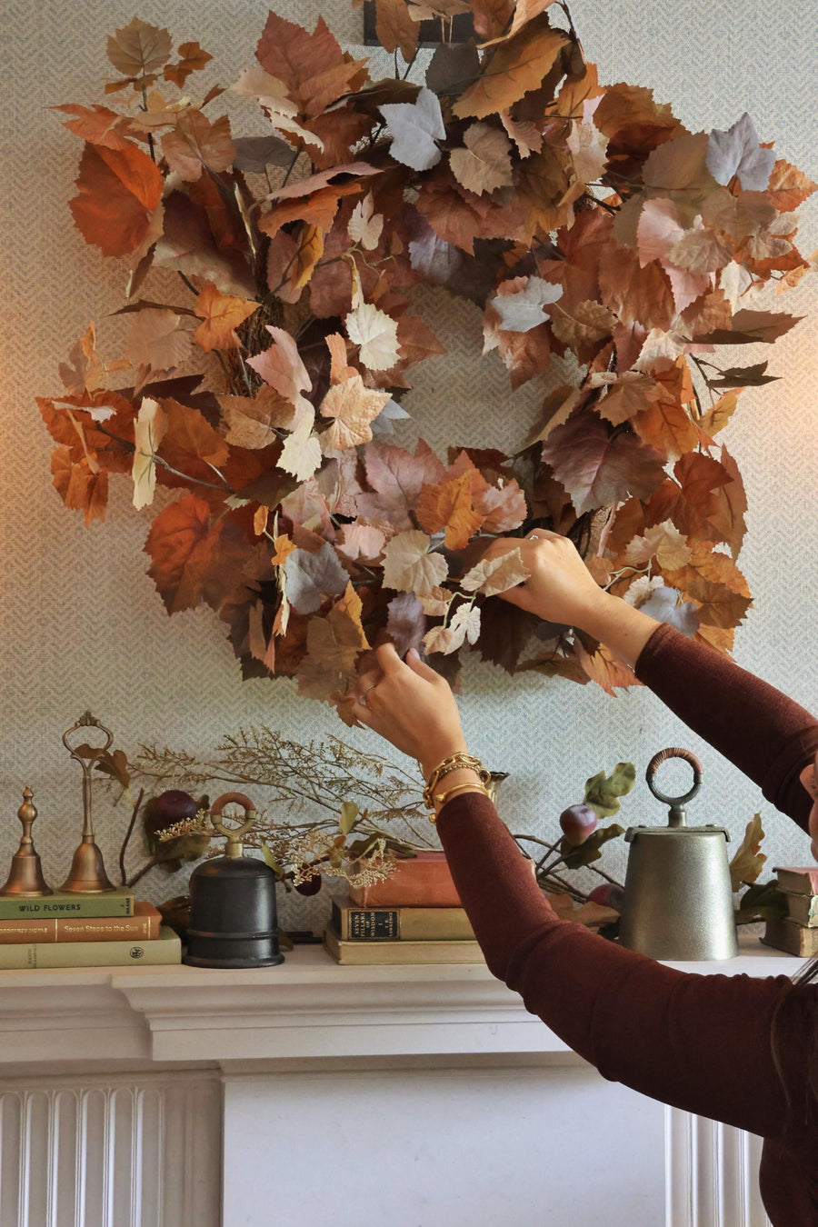 An artificial autumn leaf wreath in warm shades of brown, orange, beige, and muted blue-gray hangs on a wall above a fireplace mantel, with a person's hands adjusting the leaves.