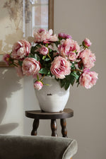 Pink flowers in a white vase on a wooden stool against a neutral wall.