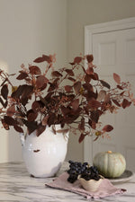 A large white ceramic vase filled with artificial autumn burgundy foliage, styled on a marble countertop next to a small green pumpkin, blackberries, and a pink cloth in a neutral indoor setting.