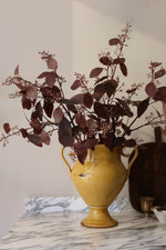A yellow ceramic vase with two handles, filled with artificial dark burgundy foliage, set on a white marble surface against a neutral beige wall.