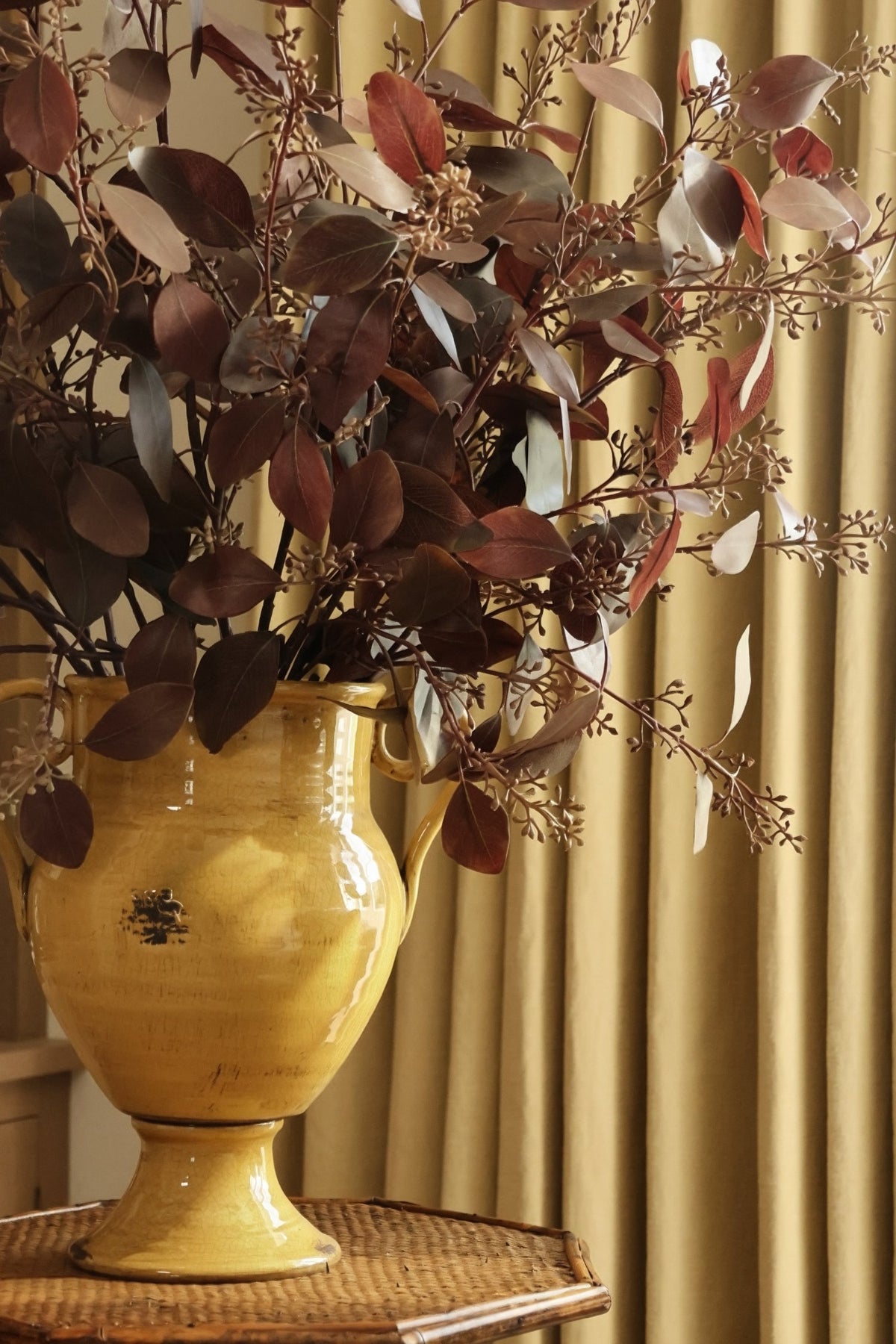 Artificial burgundy eucalyptus stems in a yellow urn vase with oak cabinetry and curtains behind 