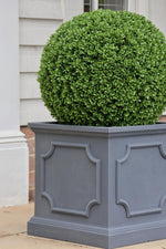 Green artificial topiary in a decorative gray planter against a light-colored wall.