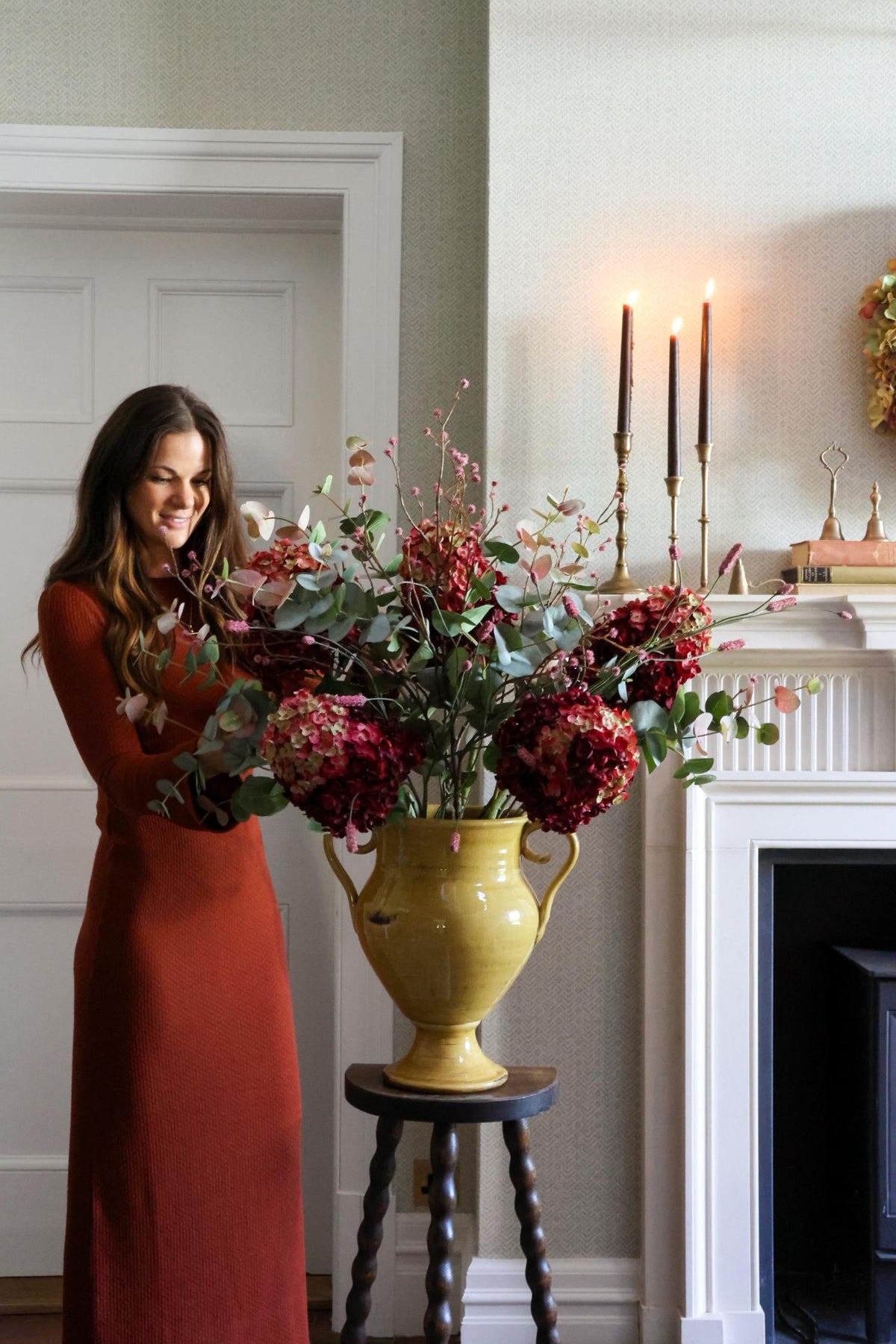 A woman in a rust-orange maxi dress stands beside a large yellow urn filled with artificial deep red hydrangeas and green foliage, placed on a small dark wooden stool in a decorated indoor room with a fireplace and lit candles.