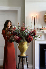 A woman in a rust-orange maxi dress stands beside a large yellow urn filled with artificial deep red hydrangeas and green foliage, placed on a small dark wooden stool in a decorated indoor room with a fireplace and lit candles.
