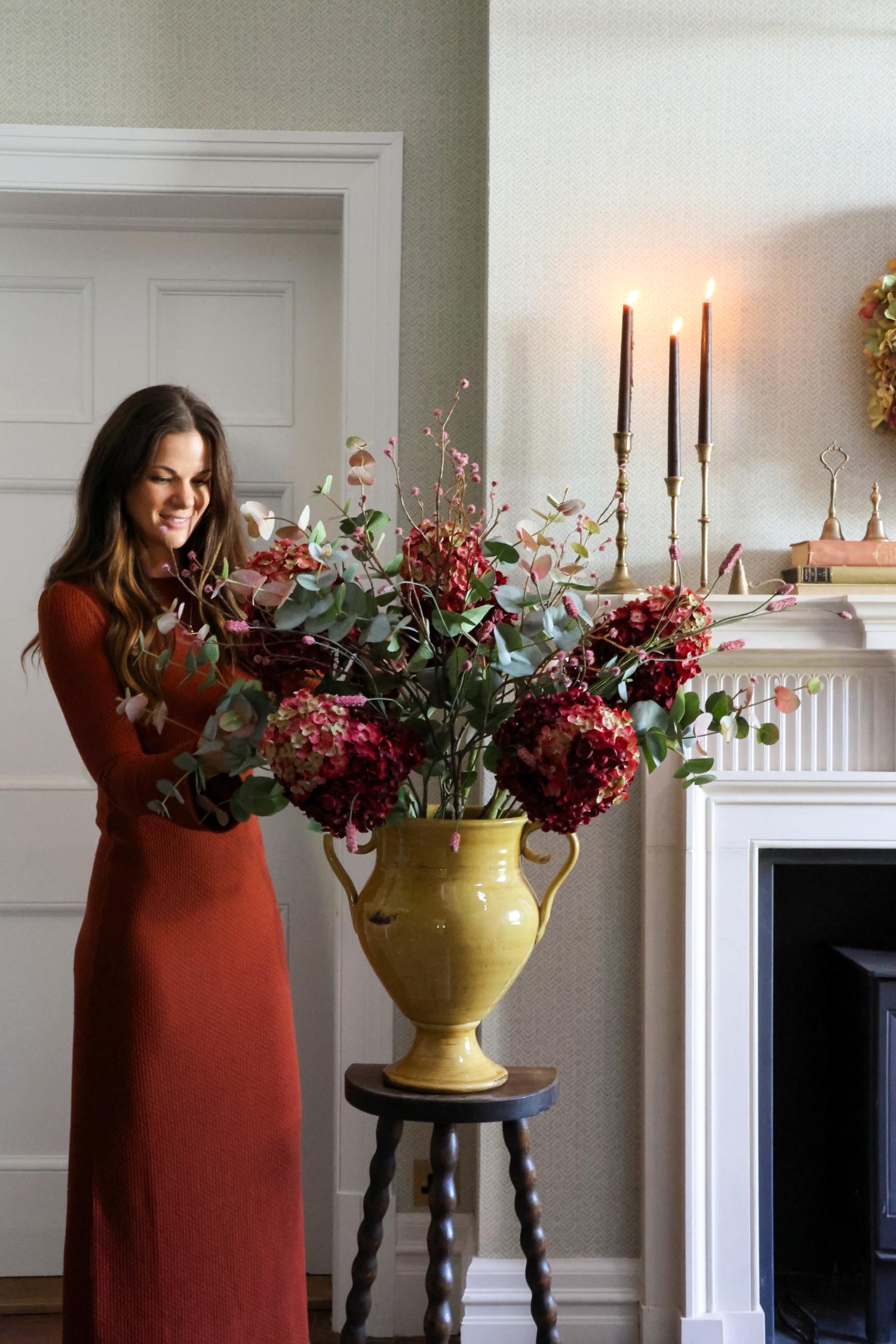 A woman in a rust-orange maxi dress stands beside a large yellow urn filled with artificial deep red hydrangeas and green foliage, placed on a small dark wooden stool in a decorated indoor room with a fireplace and lit candles.