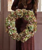 A woman in a brown velvet dress holds a large artificial dried hydrangea wreath made of green, beige, and pink petals in front of a pink door.