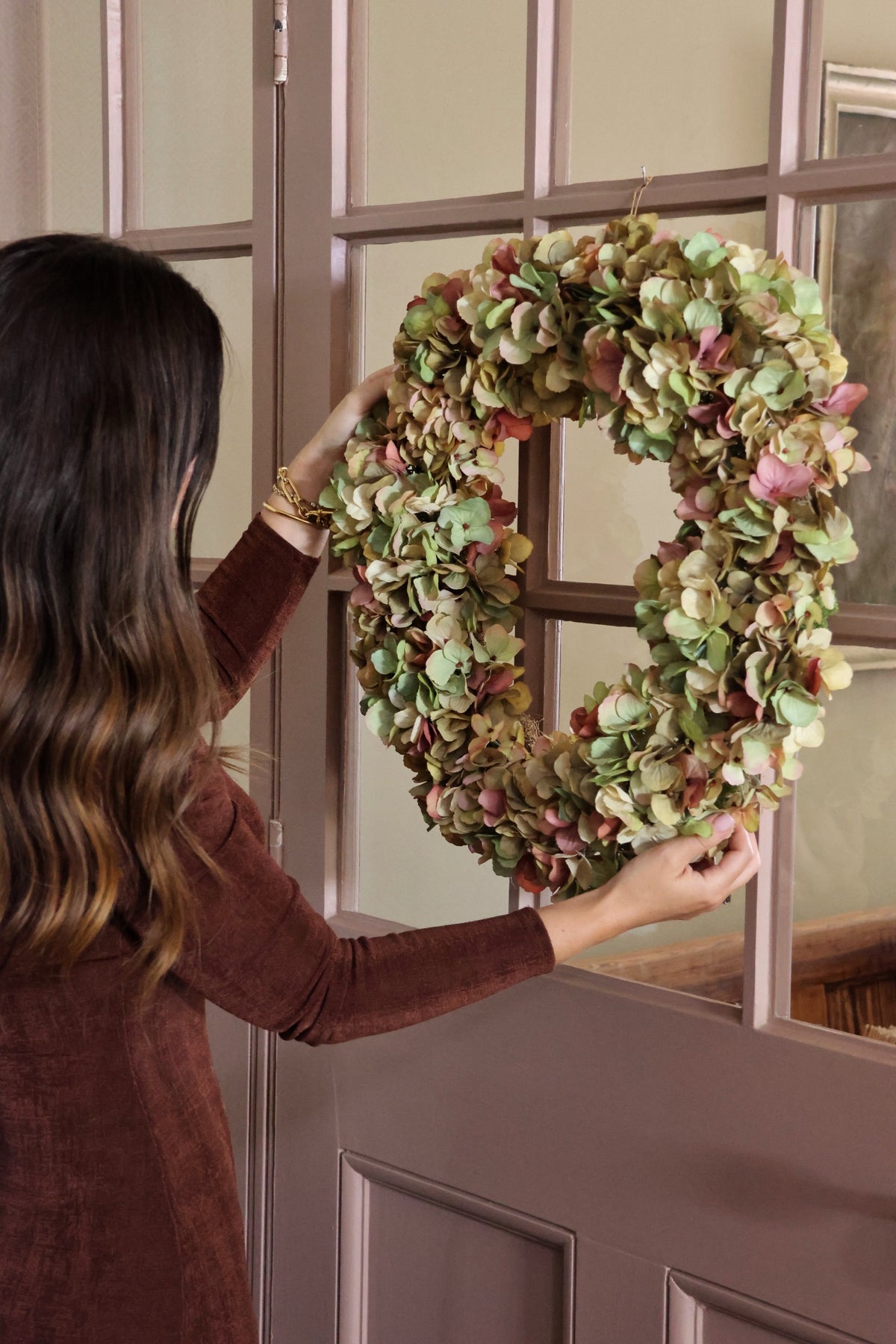 A woman in a brown long-sleeve ldress hangs a large artficial wreath made of dried green and pinkish hydrangea petals on a pink paneled door with glass panes.