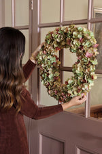 A woman in a brown long-sleeve ldress hangs a large artficial wreath made of dried green and pinkish hydrangea petals on a pink paneled door with glass panes.