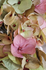 Close-up of a cluster of artificial hydrangea petals in soft pink, blush, beige, and muted green tones with small red centers.