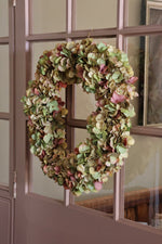 A round artificial hydrangea wreath in muted green, beige, and dusty pink tones, hanging on a brown paneled glass door.