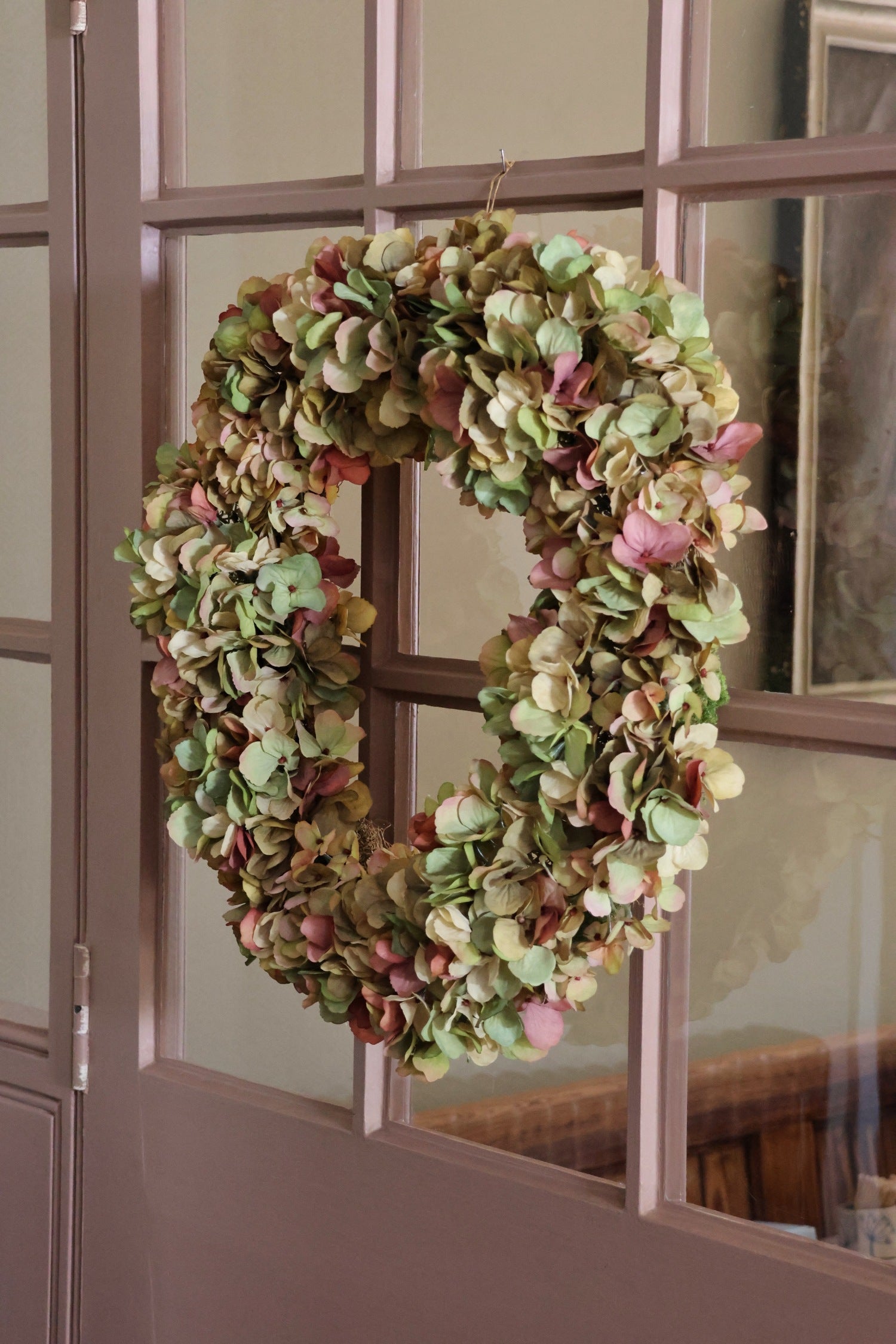 A round artificial hydrangea wreath in muted green, beige, and dusty pink tones, hanging on a brown paneled glass door.
