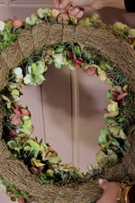 the back of an artificial wreath made on a natural brown grapevine base with faux green and pink hydrangea-style flowers and moss, with hands adjusting the top loop.