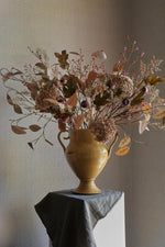 A large artificial autumn floral arrangement in a glossy mustard-yellow ceramic urn vase, displayed on a pedestal draped with a gray cloth against a neutral textured wall.