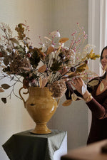 A large rustic mustarrd ceramic vase filled with an artificial floral arrangement in muted beige, brown, and dusty pink tones, styled on a draped table with a woman adjusting the arrangement in a softly lit indoor setting.