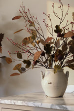 Autumn eucalyptus branch arrangement with brown leaves and small pink berries with artificial fig branches, displayed in a distressed white ceramic vase on a marble-topped surface against a neutral beige wall.