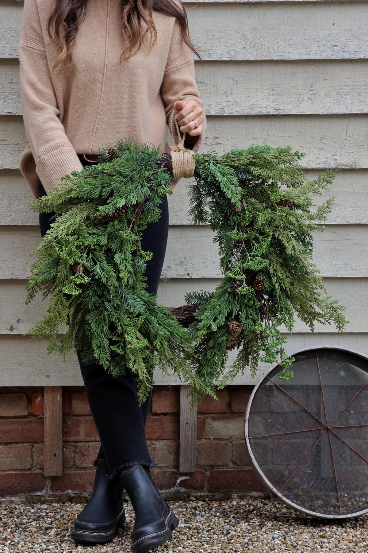 A person holding a round faux christmas wreath with green foliage and pine cones, in front of a rustic wooden backdrop with a metal wheel.