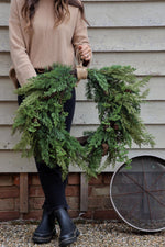 A person holding a round faux christmas wreath with green foliage and pine cones, in front of a rustic wooden backdrop with a metal wheel.