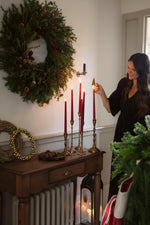 women lighting tall red candles with festive decor on a console table and large faux wreath 