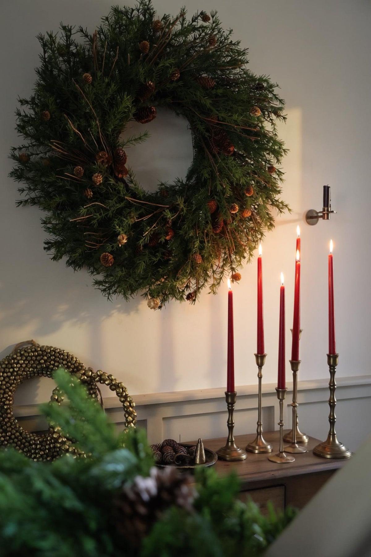 Large artificial festive wreath ontop of console table with lots of brass candlesticks and lit red candles 