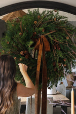women arranging branches on a large faux wreath with pinecones and larix with a brown velvet bow 