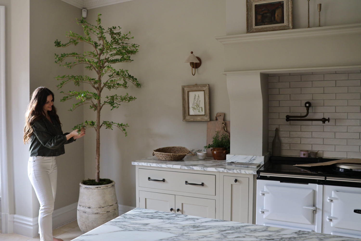 A woman in a dark green top and white pants stands next to a tall potted tree in a bright, modern kitchen with white cabinets, marble countertops, and a white subway tile backsplash.