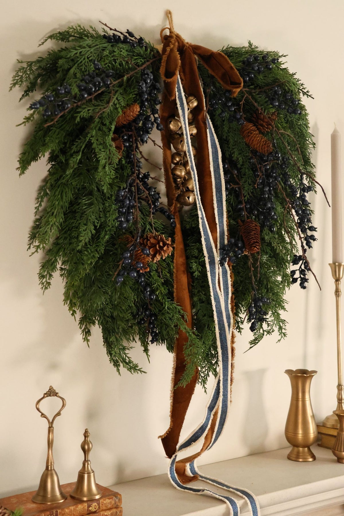 A rustic pine wreath hanging on a wall above a mantel, decorated with green pine branches, dark berries, pinecones, and a long brown velvet ribbon with a blue and white striped center stripe.