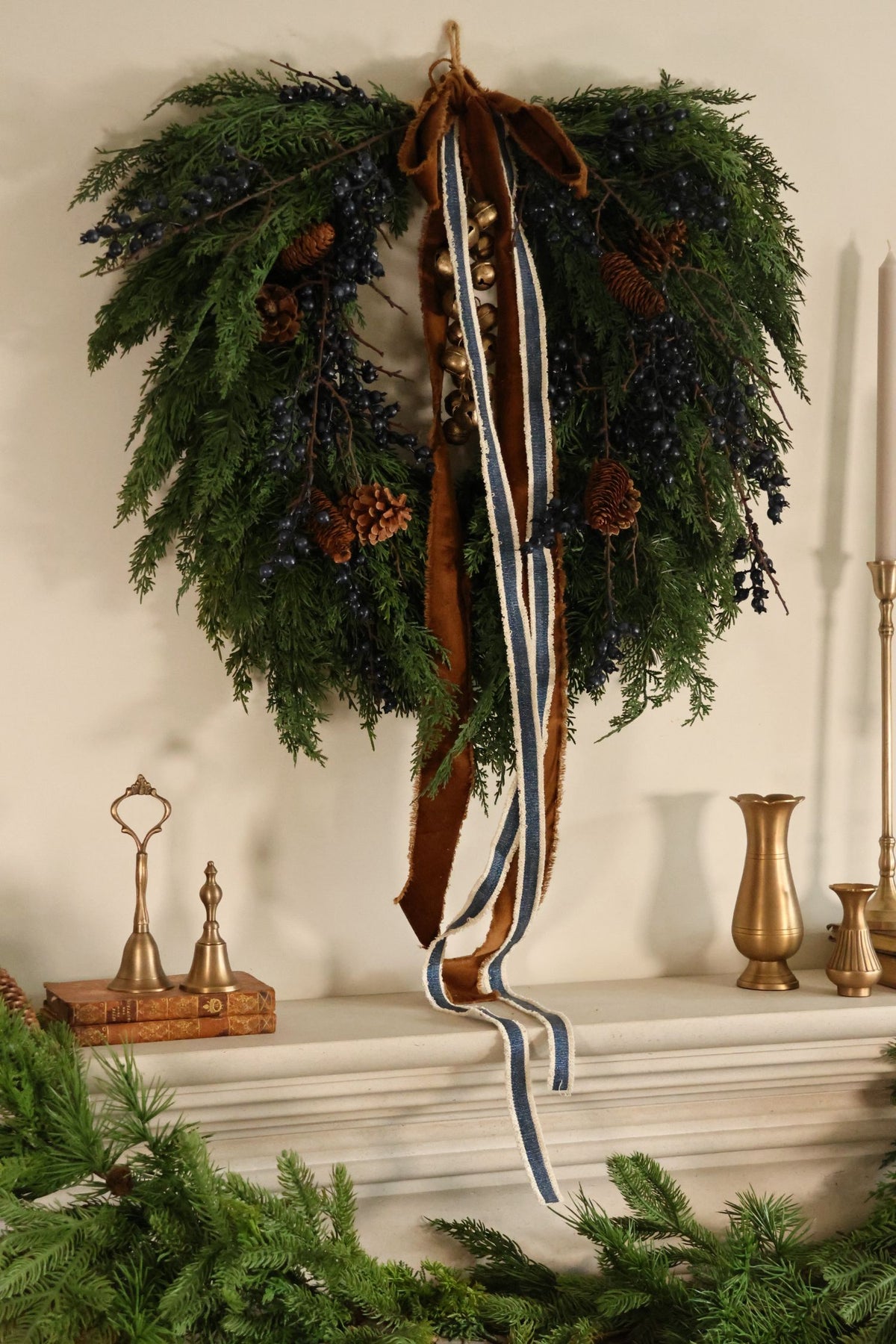 artificial juniper berry wreath with blue berries and pinecones with hanging brass bells, velvet brown ribbon and striped blue ribbon hanging above a mantel with brass bells and small bud vases 