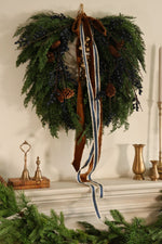 artificial juniper berry wreath with blue berries and pinecones with hanging brass bells, velvet brown ribbon and striped blue ribbon hanging above a mantel with brass bells and small bud vases 