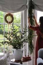 Woman arranging faux garlands and stems indoors with a large window in the background