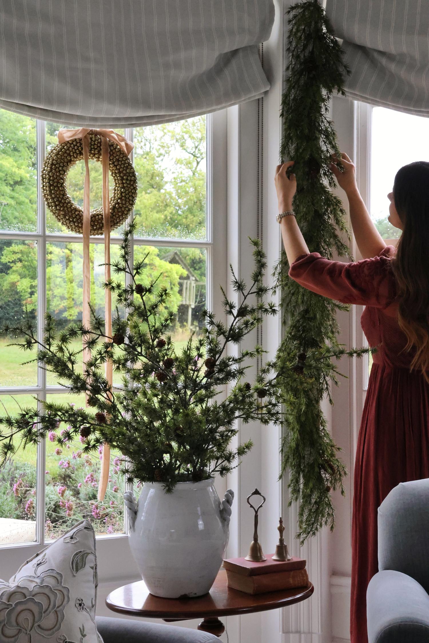 Woman arranging faux garlands and stems indoors with a large window in the background